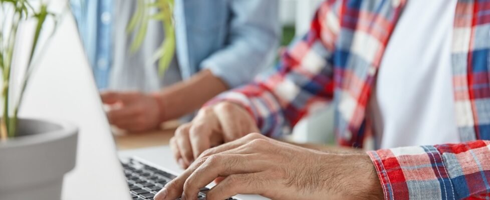 Cropped shot of two male bloggers type publication on laptop computer, use laptop computer, sit at wooden desk. Young prosperous bussinessmen check mail and send feedbacks, connected to wifi