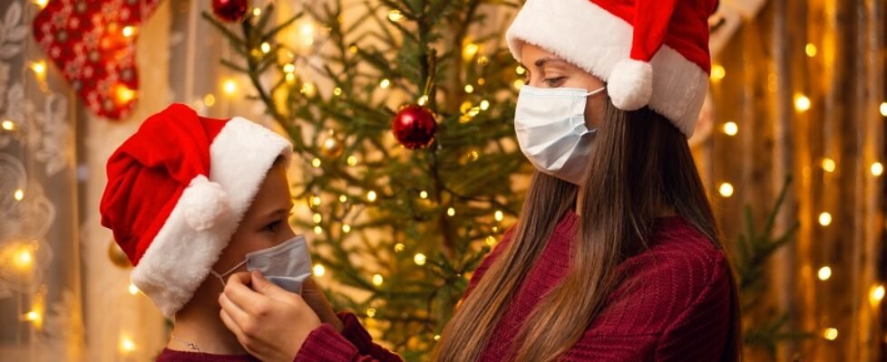 Brother and sister in christmas hats, burgundy sweaters and medical masks stand near decorated fir-tree. Family together, holidays, New Year's preparations during lockdown.