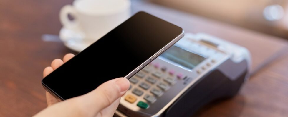 Contactless payment by phone, faceless person holding smart phone near terminal, customer paying for purchase with her cellphone, white cup with beverage on background.
