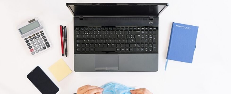 top view of a white office table with a laptop, calculator, cell phone, post it, pens, notebook and a business worker wearing a face mask