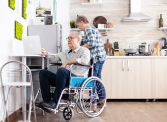 Paralysed elderly man in wheelchair typing on laptop working from home kitchen