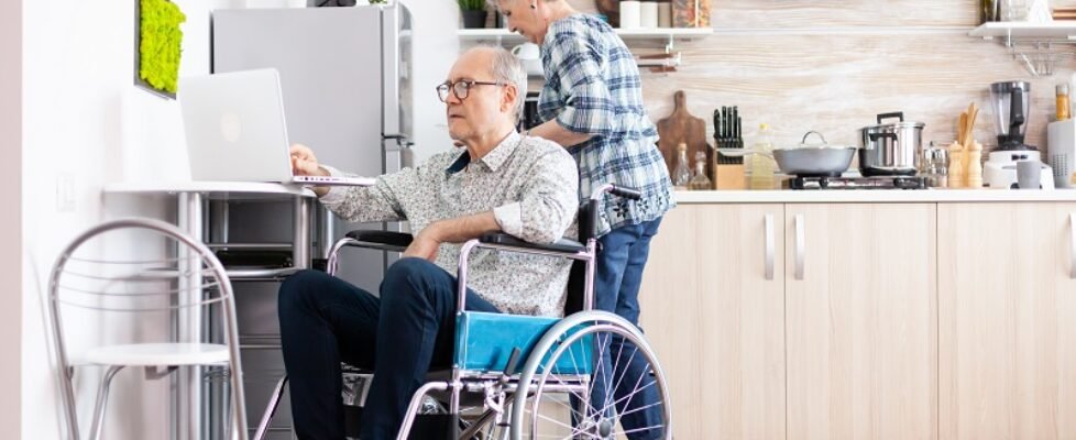 Paralysed elderly man in wheelchair typing on laptop working from home kitchen