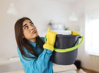 Shocked Woman Looks at the Ceiling While Collecting Water Which Leaks in the Living Room at Home. Worried Woman Holding Bucket While Water Droplets Leak From Ceiling in Living Room