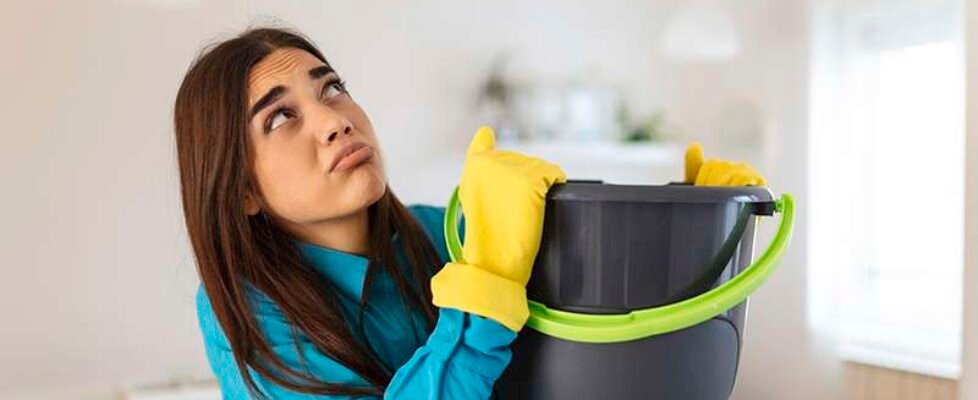 Shocked Woman Looks at the Ceiling While Collecting Water Which Leaks in the Living Room at Home. Worried Woman Holding Bucket While Water Droplets Leak From Ceiling in Living Room