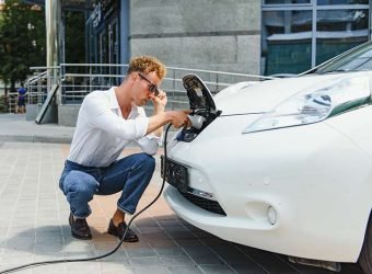 Hansome guy sitting near his new modern electric car and holding plug of the charger, while car is charging at the charging station