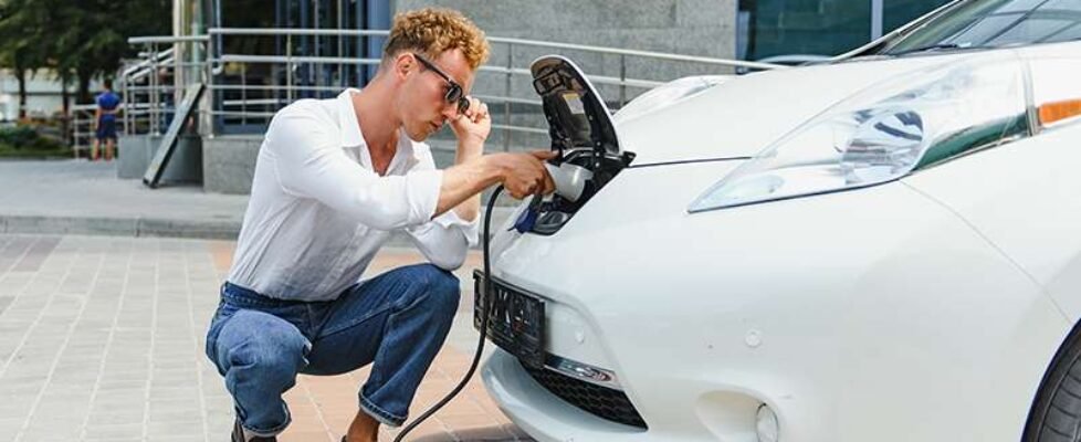 Hansome guy sitting near his new modern electric car and holding plug of the charger, while car is charging at the charging station