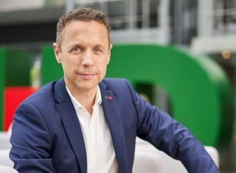 Indoor shot of serious bearded freelancer dressed in shirt, holds documentation, looks attentively at papers, does remote work, stands near window, drinks takeaway coffee. People and work concept